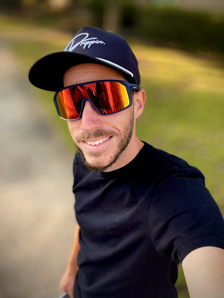 Selfie shot of Brent smiling, wearing a black shirt, a black cap and a pair of blue shades with orange lenses.